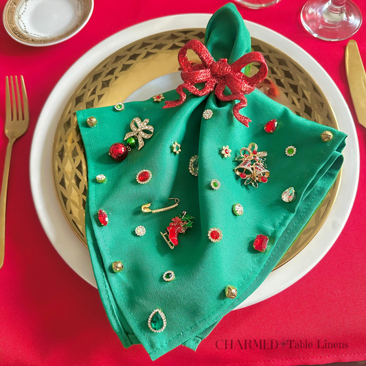 Charmed Table Linens - Napkins are a beautiful bright holiday green with a red glittery bow, embellished with brooches (holiday bells, red ice skating boot, and a red ornament charm, gems, and charms of red, green and diamon like charms. 